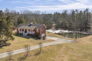 A red house with white trim sits on a grassy lot near a small pond with a dock at 8437 Charlie Stovall Road, Oxford. A gravel driveway leads to the house, surrounded by trees and open land under a blue sky.