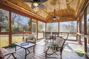 A screened-in porch with a wooden ceiling and floor, containing cushioned chairs, a coffee table, a high-top dining table with stools, ceiling fans, and views of a grassy yard and trees outside.