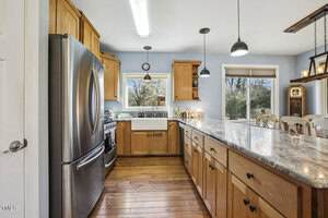 Modern kitchen with stainless steel appliances, wooden cabinets, farmhouse sink, marble countertop, pendant lights, and large windows overlooking trees; dining area visible in background.