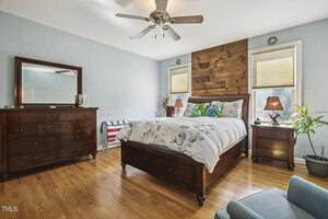 A bedroom with light blue walls, a wooden bed with floral bedding, matching nightstands and dresser, a ceiling fan, hardwood floors, two windows with blinds, and a potted plant by the window.