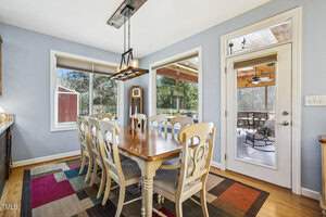 A dining area with a wooden table and eight chairs on a colorful rug, next to large windows and a glass door leading to a covered porch with outdoor seating. Natural light fills the space.