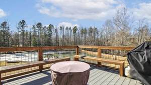A wooden deck with a railing overlooks a pond and wooded area. There is a covered round table, a bench, and a grill on the deck. The sky is partly cloudy and the trees are mostly bare.