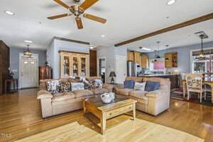 A spacious open-concept living area features a beige sectional sofa, wooden coffee table, ceiling fans, and hardwood floors. The space flows into a kitchen with wood cabinets and a dining area with a rectangular table.
