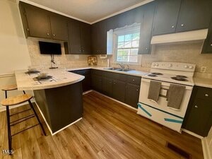 A kitchen with dark cabinets, white countertops, a white electric stove, a double sink below a window, and a small breakfast bar with two wooden stools. The floor is wood-patterned, and there is a wall-mounted TV.