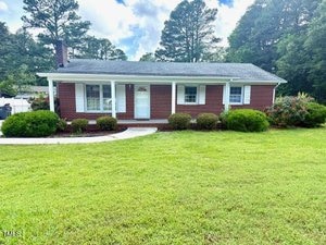 A one-story red brick house with white trim, a covered front porch, and a neatly maintained lawn with shrubs in front. Tall trees and a partly cloudy sky are visible in the background.