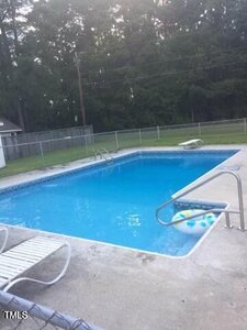 A rectangular outdoor swimming pool with clear blue water, a diving board, and a metal handrail. There are pool floats in the water and lounge chairs on the concrete deck. A chain-link fence and trees are in the background.