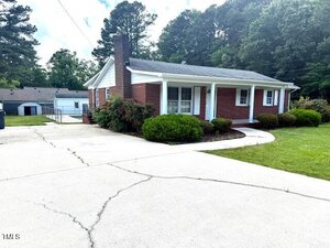 A charming one-story brick house at 100 E Dale Drive, Oxford, features white columns and a covered front porch, surrounded by bushes and a lawn. A long concrete driveway leads to the home, set against a scenic backdrop of trees.
