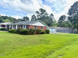 Single-story red brick house with white trim and a covered front porch, surrounded by green lawn and bushes. A detached garage and fenced backyard are visible. The sky is partly cloudy with trees in the background.