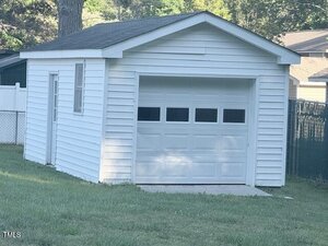 A small white detached garage with a single garage door and a side door, featuring horizontal siding and a shingled roof, situated on a grassy lawn with a fence in the background.
