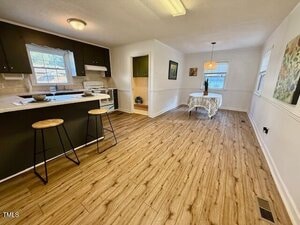 A kitchen with dark cabinets, white appliances, and a breakfast bar with two stools is shown. The kitchen opens into a dining area with a small round table and chairs by a window. The floor is wood-patterned.