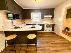 A kitchen with dark brown cabinets, a white stove and range hood, a window above the sink, a light-colored countertop with two place settings, and two wooden stools on a wood floor. A small TV is mounted under a cabinet.