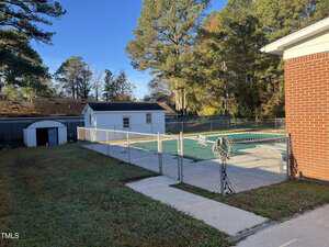 A fenced-in outdoor swimming pool with a green cover next to a brick building, a concrete walkway, grass, a white shed, and trees in the background under a clear blue sky.