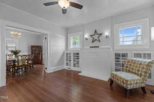 A bright living room with hardwood floors, white walls, a ceiling fan, built-in cabinets with glass doors, a patterned armchair, and a white brick fireplace. A dining area is visible through an open doorway.