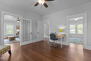 A home office with a wooden floor, a desk and chair, ceiling fan, and white walls. Two adjoining rooms are visible, each with large windows and ceiling fans, and a patterned chair sits in the foreground.