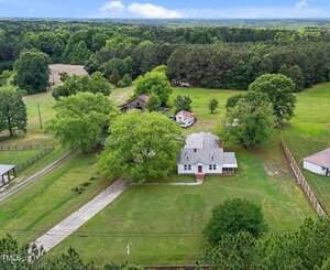 Aerial view of a white two-story house with a gray roof, a red front door, and a long driveway, surrounded by trees, grassy fields, and fenced areas under a cloudy sky.