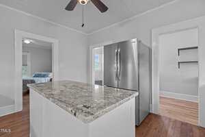 A modern kitchen with a marble island countertop, stainless steel refrigerator, and hardwood floors. Doorways lead to a bedroom, a room with shelves, and another area with a window.