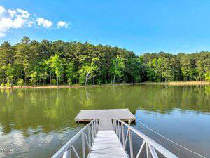 A metal dock with a wooden platform extends over a calm lake on the 1.65 acres Buchanan Road, Manson, surrounded by dense green trees under a clear blue sky.