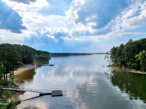 A calm lake bordered by dense trees and sandy beaches, with a dock extending into the water and clouds scattered across a bright sky.