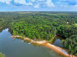 Aerial view of a wooded peninsula surrounded by a lake, with several docks extending into the water and a sandy shoreline. Dense forest covers most of the land, and the sky is partly cloudy.