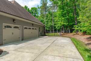 A concrete driveway leads to a two-car garage attached to a gray house. The garage doors have decorative windows, and the area is bordered by green grass, trees, and a black metal fence.
