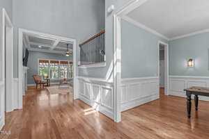 A hallway with light blue walls, white wainscoting, and wood floors leads to an open living area with large windows, a ceiling fan, and a wooden chair. Natural light enters through the windows.