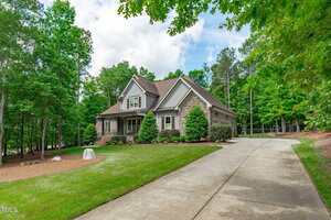 A two-story house with a stone and siding exterior, front porch, and gabled roof sits beside a long curved driveway, surrounded by green lawns, trees, and landscaped shrubs under a partly cloudy sky.