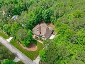 Aerial view of a two-story house with a brown roof, surrounded by trees and greenery, located in a suburban neighborhood with nearby houses and a paved road.