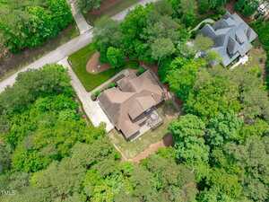 Aerial view of a suburban house with a brown roof, driveway, and surrounding green trees. A road runs in front of the property, and a neighboring house is visible to the right.