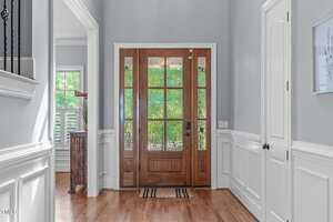 A bright entryway with light gray walls, white wainscoting, and a wooden front door with glass panels. Natural light enters through the door, and a small striped rug is placed on the wood floor.
