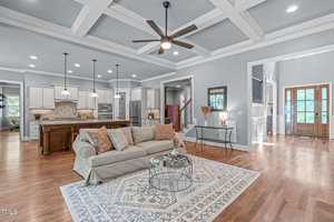 Spacious open-concept living area with a beige sofa and patterned rug, coffered ceiling, ceiling fan, and view of the kitchen with white cabinets, island, pendant lights, and entryway with wooden double doors.