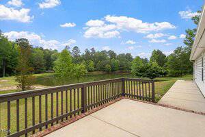 A spacious patio with a wooden railing overlooks a pond surrounded by grass, trees, and forest under a blue sky with scattered clouds. Part of a white house and a concrete walkway are visible on the right side.