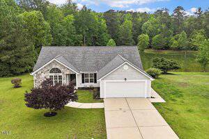 Single-story house with gray siding and stone accent, attached two-car garage, driveway, and a well-kept lawn. Surrounded by trees and located near a pond with forest in the background.