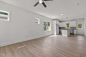 A modern open-concept living area with light wood flooring, white walls, three square windows, ceiling fan, and a view into a white kitchen with stainless steel appliances and an island.