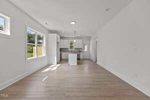 A bright, empty room with light wood floors and white walls leads to a modern kitchen area with white cabinets, an island, and stainless steel appliances. Large windows provide natural light and a view of the outdoors.