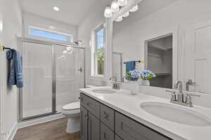 A modern bathroom with a double-sink vanity, gray cabinets, a large mirror, a glass shower enclosure, a toilet, white walls, wood flooring, and a potted flower arrangement on the counter.