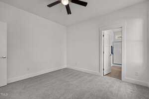 A small, empty room with white walls, gray carpet, a ceiling fan with dark blades, and an open door leading to a bright hallway with wood flooring.