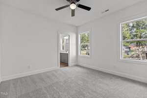 A bright, empty bedroom with light gray carpet, white walls, a ceiling fan with light, two windows, and a doorway leading to a bathroom with a double-sink vanity and outdoor views.
