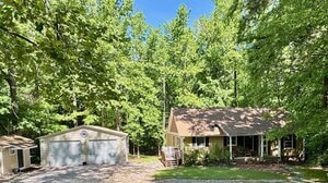 A single-story tan house with a front porch sits among tall green trees. To the left is a detached two-car garage with light-colored doors. A gravel driveway and lawn are in the foreground under a clear blue sky.
