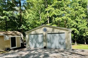 A tan double garage with two closed metal roll-up doors and a blue compass decoration above them, situated on a gravel driveway and surrounded by tall green trees. A smaller tan shed is visible to the left.