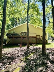 A yellow single-story house with a raised wooden deck, surrounded by tall trees and a shaded yard with patches of sunlight and grass. Wooden benches are placed in the yard.