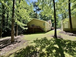 A tan single-story house with a wooden deck is surrounded by tall trees and green grass. There is a small outbuilding to the right and sunlight filters through the leafy canopy overhead.