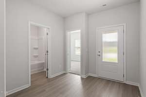 A hallway with light wood flooring, white walls, a closed frosted glass door, an open door leading to a carpeted room with a window, and another open door showing a bathroom with a tub and shelves.