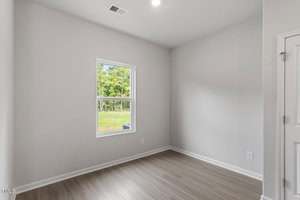 A small, empty room with light gray walls, a single window showing greenery outside, and light wood flooring. There is a white door to the right, and the room is lit by a ceiling light.