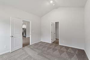A carpeted bedroom with light gray walls, a vaulted ceiling, and two white doors: one open to a hallway with a glimpse of a kitchen, and another leading to a small interior room.