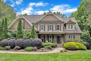 This large two-story house at 3007 Krogen Court in Creedmoor features a brick and siding exterior, multiple gables, a covered front porch, and well-maintained landscaping with bushes, shrubs, and trees under an overcast sky with scattered clouds.