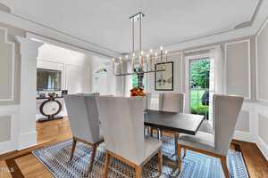 A dining room with a rectangular table, six high-back chairs, a modern chandelier, a wall mirror, artwork, large windows, and hardwood floors with a patterned rug.