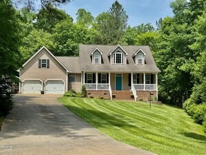 A beige two-story house at 327 Beavertail Road with a front porch, three dormer windows, a teal front door, and an attached two-car garage sits at the end of a curved driveway in Henderson, surrounded by lawn and dense green trees.
