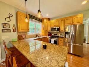 A kitchen with wooden cabinets, stainless steel appliances, granite countertops, and two pendant lights. There are two chairs at the counter and a window above the sink letting in natural light.
