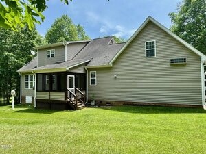A beige two-story house with a dark roof, screened porch, and wooden steps leading to a grassy backyard surrounded by trees under a blue sky.