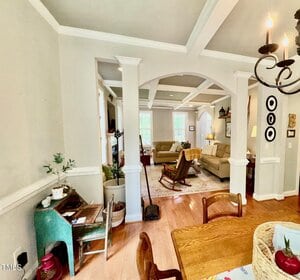 A view from a dining area into a living room with arched openings and white columns, wood floors, beige walls, and coffered ceilings. There are various chairs, a sofa, and decor such as plants and wall art.